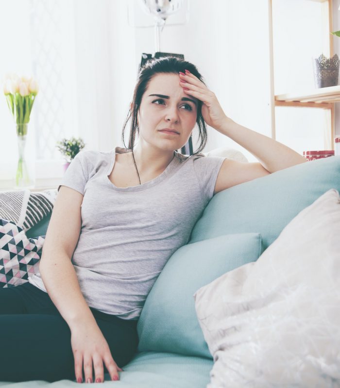 Young worried woman with headache while sitting on comfortable sofa